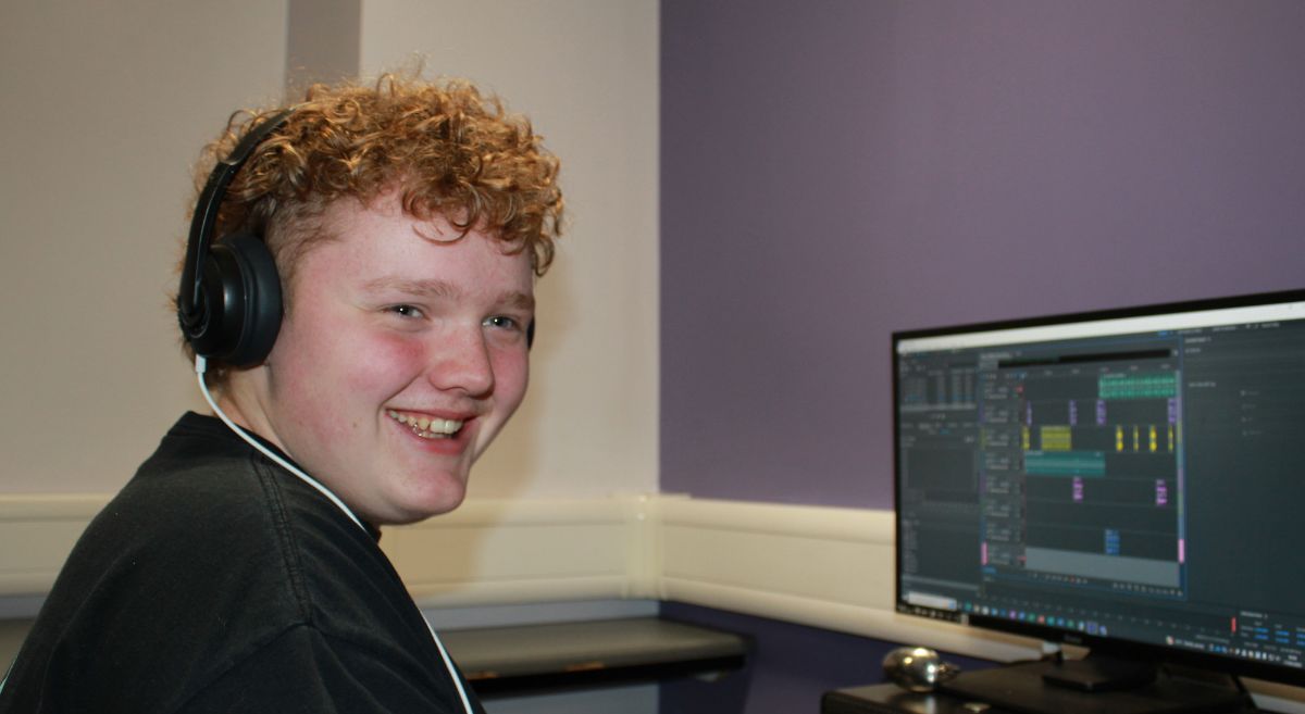 Young man with dark curly hair, wearing a black teeshirt and headphones, smiling to camera and pictured at at computer screen.